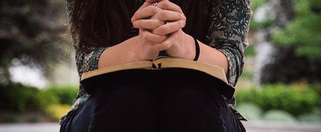 Person sitting down praying with her hands on the bible
